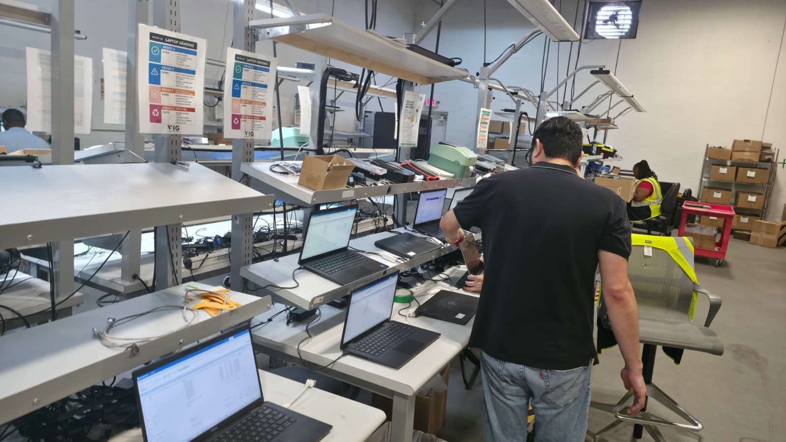 A VIG technician grading laptops at the audit line beneath the posted grading rubric
